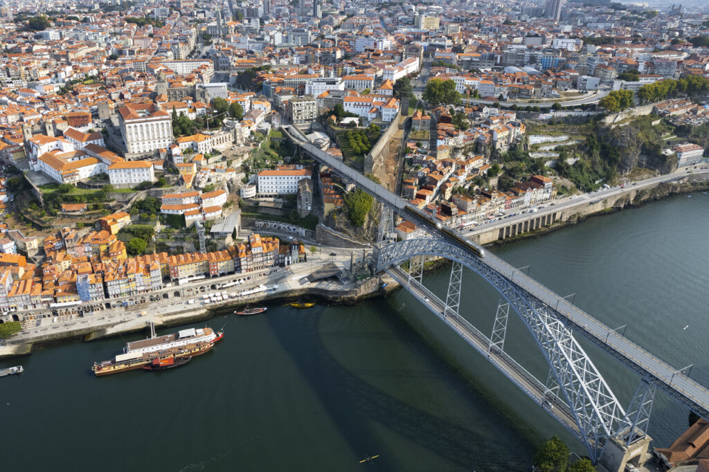 Panoramic aerial view of Lisbon cityscape with traditional orange rooftops, the Tagus River, and the 25 de Abril Bridge in the distance.