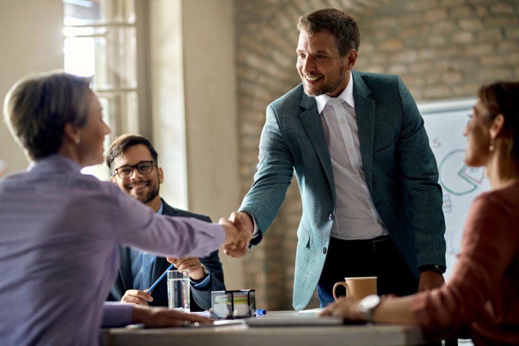 A male real estate advisor smiling and shaking hands with a client during a business meeting in an office setting.